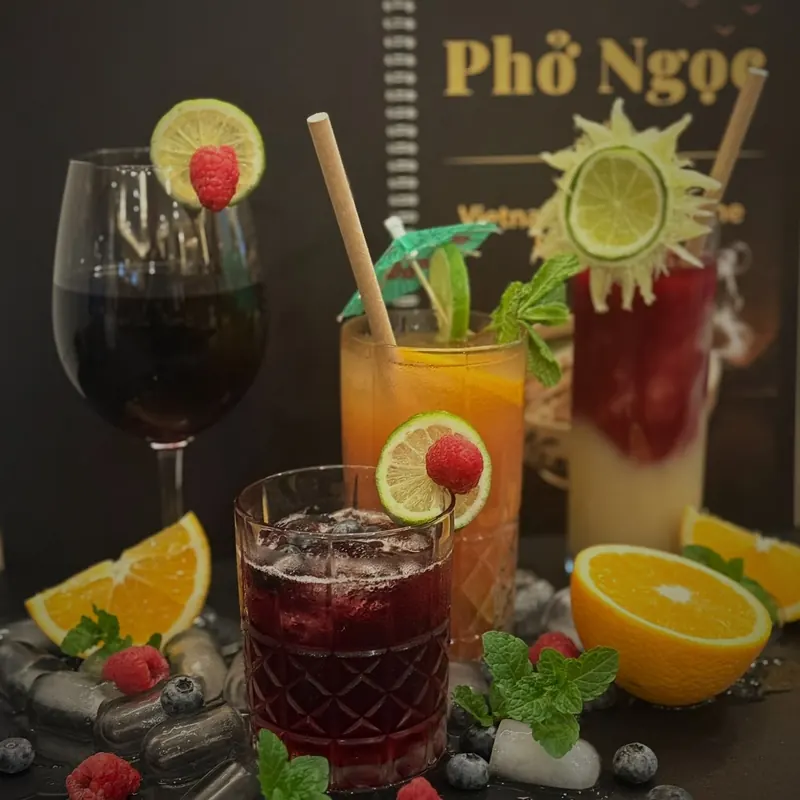 Vier Getränke stehen auf einer glänzenden schwarzen Fläche vor einer aufgestellten dunklen Karte mit dem goldenen Schriftzug „Phở Ngọc“: ein Weinglas und ein kurzes geschliffenes Glas mit dunkelrotem Getränk, beide mit Limettenscheibe und Himbeere, ein hohes Glas mit orangefarbenem Getränk und grünem Papierschirmchen sowie ein hohes Glas mit roter und hellgelber Schicht und sonnenförmiger Limettendeko, umgeben von Orangen, Himbeeren, Heidelbeeren, Minze und Eiswürfeln.