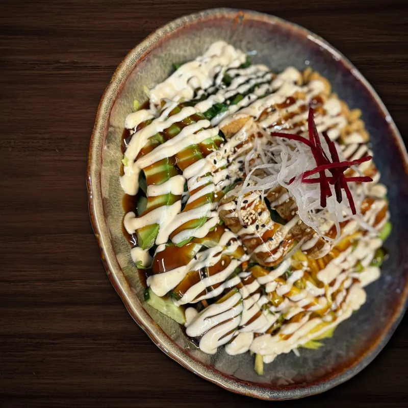 Große Schale mit Avocado, Gurke, Mango und frittierten Stücken, überzogen mit dunkler und heller Soße, bestreut mit Sesam und garniert mit Rettich- und Rote-Bete-Streifen.