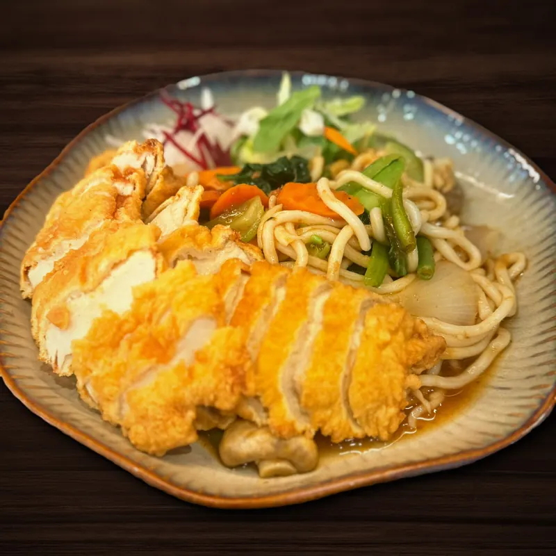Teller mit dicken Nudeln und gebratenem Gemüse wie Karotten, Pak Choi, grünen Bohnen und Zwiebeln, darauf ein in Scheiben geschnittenes, goldbraun frittiertes Hähnchenstück.