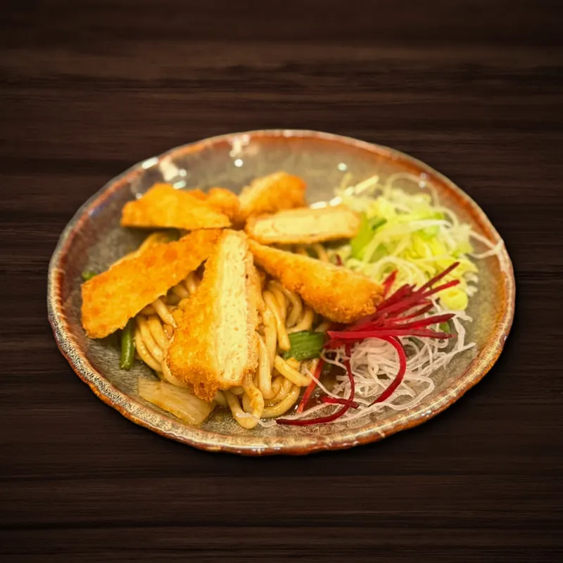 Schale mit dicken Nudeln in dunkler Soße mit grünen Bohnen und Zwiebeln, darauf fächerförmig angeordnete panierte, frittierte Streifen, dazu Salat mit Rettich- und Rote-Bete-Streifen.