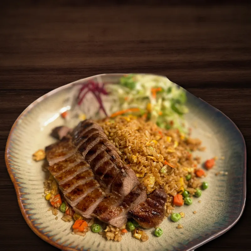 Mehrere Scheiben gegrilltes Fleisch mit Grillstreifen liegen auf gebratenem Reis mit Erbsen, Karottenwürfeln, Mais und Ei, dahinter fein geschnittener Kohl mit Karotten- und violetten Gemüsestreifen, auf einem Keramikteller mit gewelltem Rand.