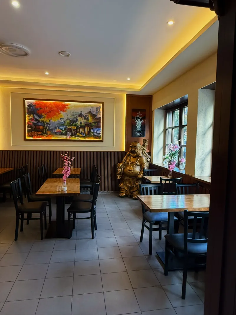 Eine Ecke des Gastraums mit Holztischen und Blumenvasen, einem farbigen Gemälde eines vietnamesischen Dorfes, einer goldenen lachenden Buddha-Figur und Fenstern mit Orchideen.