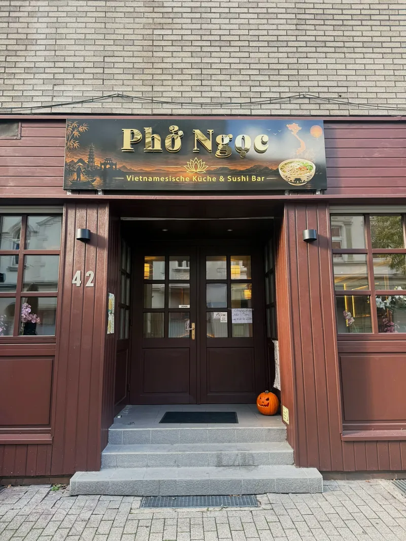 Der Eingang des Phở Ngọc an der Essener Straße 42: dunkelrote Holzfassade mit beleuchtetem Schild „Phở Ngọc, Vietnamesische Küche & Sushi Bar“, darunter eine zweiflügelige Holztür mit Sprossenfenstern und drei Stufen.